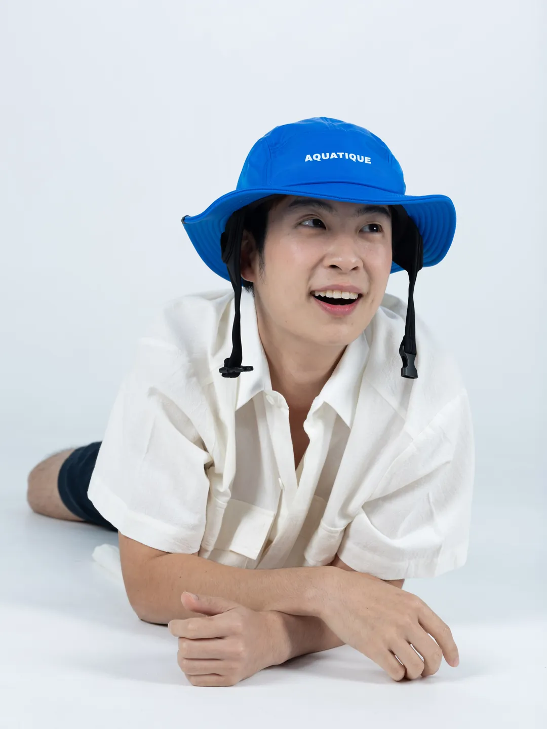A man lying on his front and smiling, wearing a bright blue AQUATIQUE bucket hat
