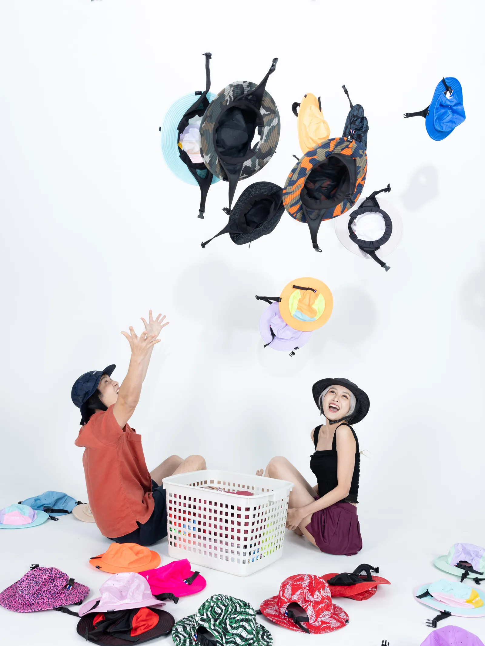 Two people sitting on a white studio floor, throwing AQUATIQUE hats in many colours into the air, more hats and a basket around them