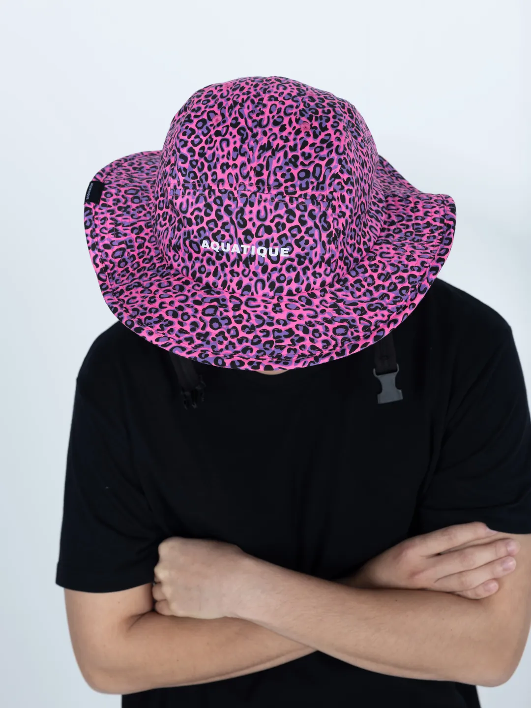 A person with arms crossed, a pink leopard-print AQUATIQUE bucket hat pulled down over the face