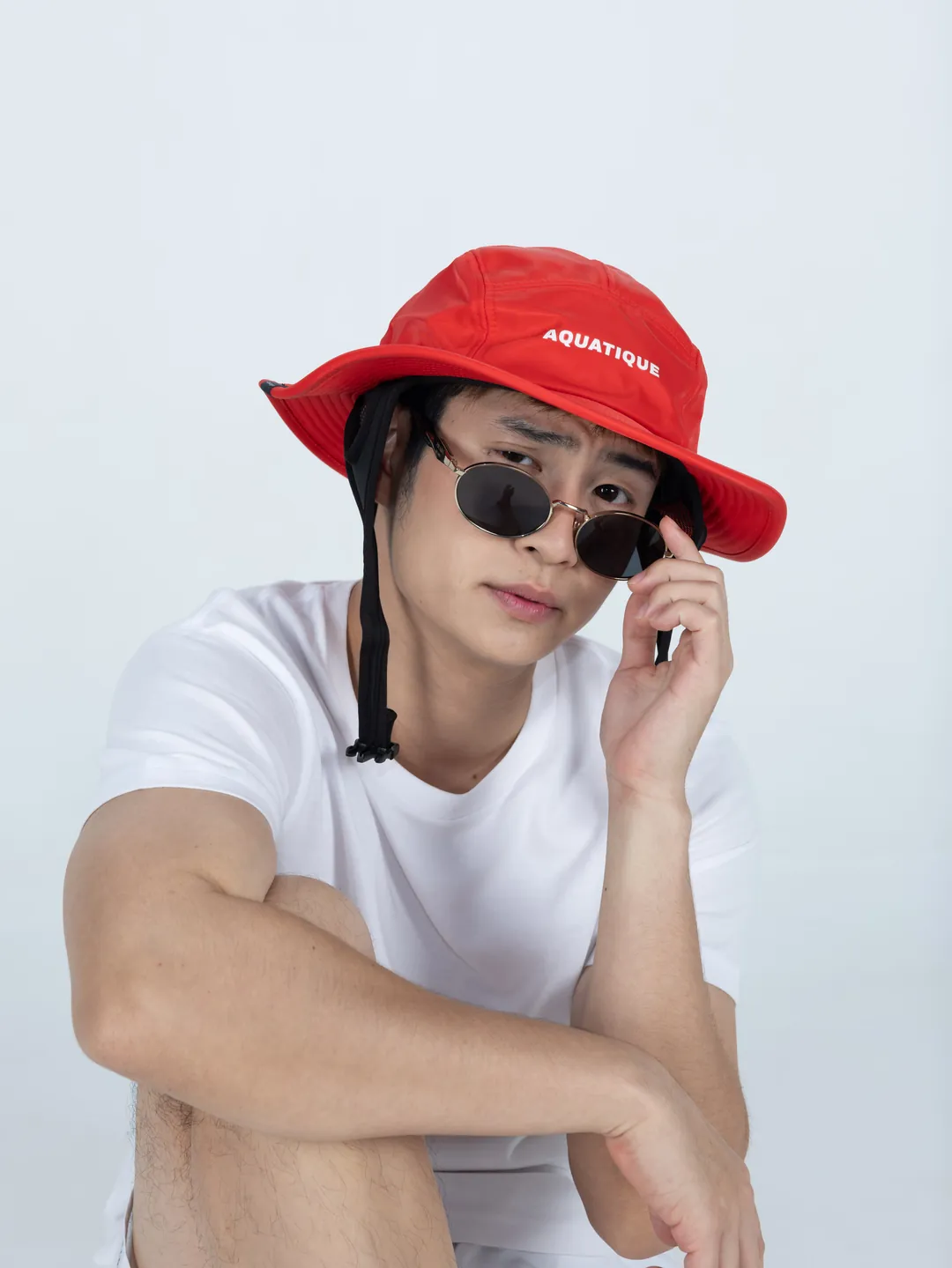 A man crouching in a red AQUATIQUE bucket hat and sunglasses, the logo on the front of the hat