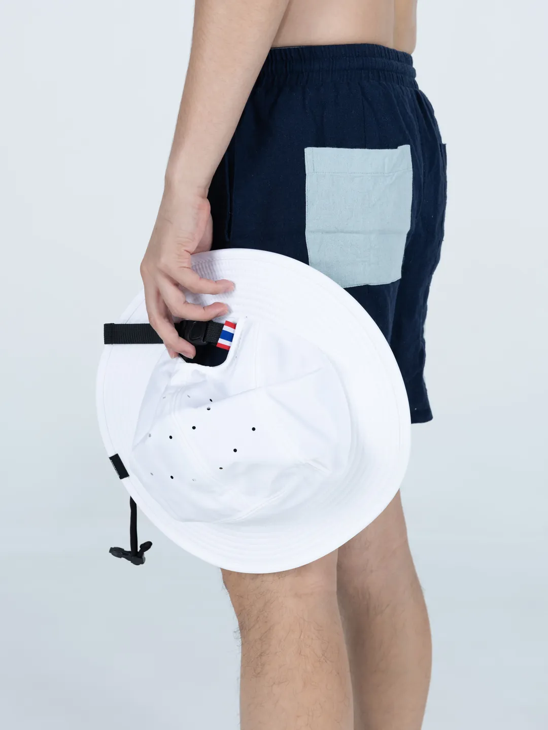 A white AQUATIQUE bucket hat held at the hip, showing the vent holes, the chin cord toggle and a Thai flag tab